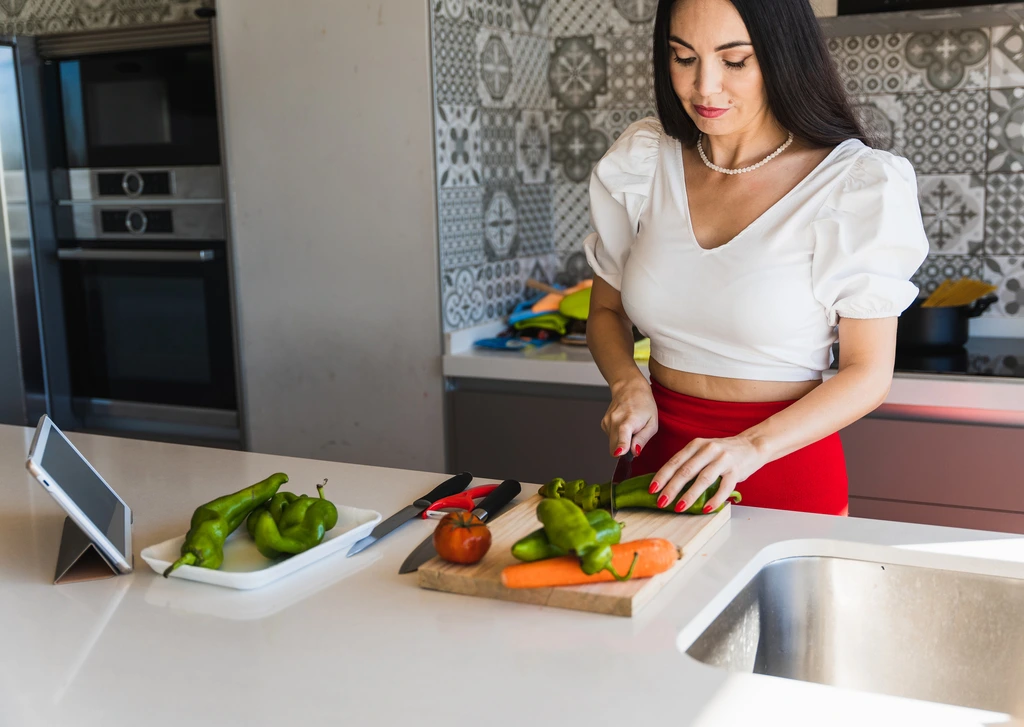 Felicia 'Felix' Hayes chopping green peppers on a wooden cutting board in her modern kitchen while looking at a recipe on a tablet.