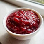 A small white bowl filled with vibrant, thick homemade raspberry jam, showing visible seeds.