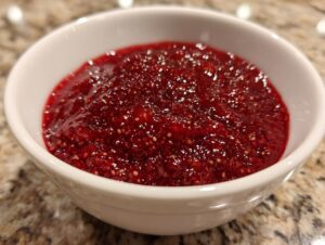 A close-up shot of thick, glistening, deep red raspberry jam filled in a small white bowl.