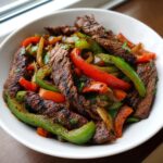 Close-up of perfectly cooked steak fajitas strips mixed with vibrant red, green, and orange bell peppers and onions in a white bowl.