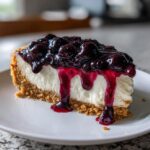 A decadent slice of blueberry cheesecake with a thick graham cracker crust and rich blueberry topping dripping down the side.