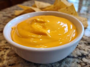 Close-up of a small white bowl filled with smooth, bright orange nacho cheese dip, with tortilla chips blurred in the background.