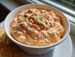 A close-up of creamy, orange-pink smoked salmon dip garnished with fresh dill, served with crackers.