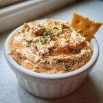 Close-up of creamy, orange-hued smoked salmon dip topped with fresh dill, served in a white bowl with a cracker.