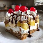 A decadent slice of banana split cake featuring layers of crust, cream, pineapple, banana, topped with whipped cream, chocolate syrup, and maraschino cherries.