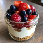 Close-up of a single serving Protein Cheesecake layered in a glass jar with a graham cracker crust, creamy filling, and fresh mixed berries.