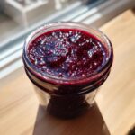 Close-up of a glass jar filled with rich, dark purple blackberry jam, glistening in the sunlight.