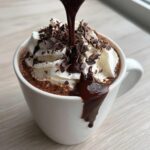Close-up of boozy hot chocolate topped with whipped cream, chocolate shavings, and rich chocolate syrup being poured over it.