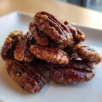 Close-up of shiny, caramelized candied pecans piled on a white plate, showcasing the sugary coating.