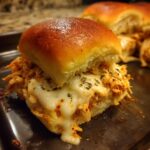 A close-up of a gooey, cheesy buffalo chicken sliders sandwich on a baking sheet, topped with herbs.