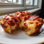 A close-up of a pepperoni pizza muffin with melted, stretching cheese, showing a bite taken out of the fluffy base.
