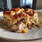 A close-up serving of chicken pot pie casserole topped with golden biscuits, showing creamy filling and mixed vegetables.