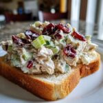 Close-up of a generous scoop of chicken salad sandwich mix, featuring chicken, celery, and dried cranberries, piled on a slice of toasted bread.