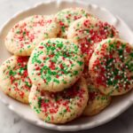 A close-up of several soft, round christmas sprinkle sugar cookies topped with red, green, and white nonpareils on a white plate.