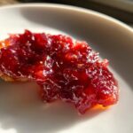 A close-up, glossy spoonful of deep red christmas jam spread on a piece of toast or cracker, sitting on a white plate.