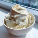 A generous serving of creamy, pale yellow banana ice cream topped with a sprinkle of spice in a white bowl.