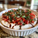 A white, fluted dish filled with creamy bruschetta dip, topped with diced tomatoes, fresh basil, and balsamic glaze.