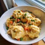 Close-up of creamy chicken and dumplings in a white bowl, garnished with peas, carrots, and parsley.