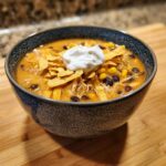 A close-up of a bowl of creamy chicken tortilla soup topped with tortilla strips, black beans, corn, and sour cream.