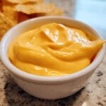 Close-up of smooth, bright orange nacho cheese dip in a small white bowl, with tortilla chips blurred in the background.