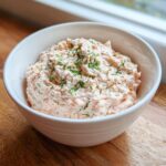 A white bowl filled with creamy, pink smoked salmon dip, garnished generously with fresh dill.