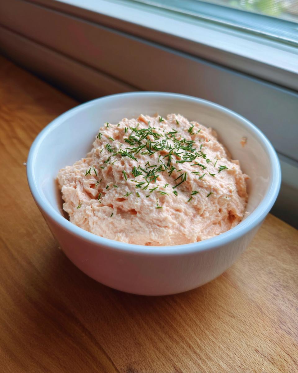 A white bowl filled with creamy, pink smoked salmon dip, topped with fresh dill.