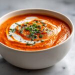 A close-up of a vibrant orange tomato bisque, swirled with cream and topped with fresh herbs and pepper.