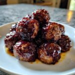 A close-up of several shiny Crockpot grape jelly meatballs piled high on a white serving plate.