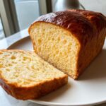 Close-up of a loaf of golden brown, freshly baked brioche bread, sliced to show the soft, yellow interior crumb.