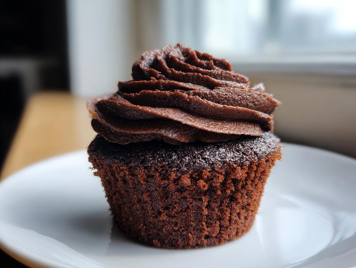 A close-up of one moist, dark chocolate cupcake topped with perfectly piped chocolate frosting, ready to eat.