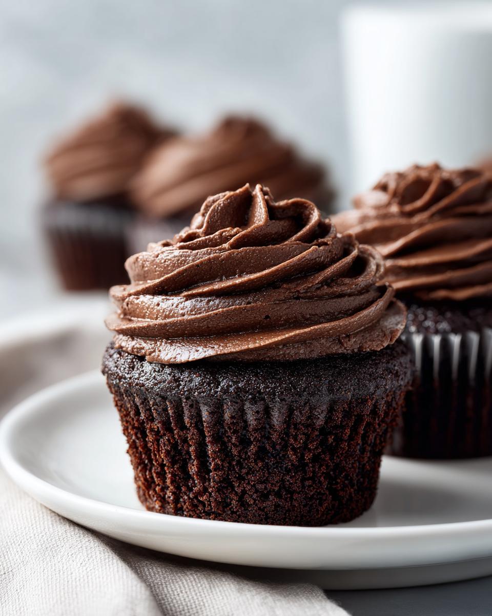 Close-up of one of the easy chocolate cupcakes topped with thick, swirled chocolate frosting on a white plate.