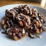 A pile of finished crockpot peanut clusters coated in dark chocolate and drizzled with white chocolate on a white plate.