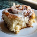 A close-up, mouth-watering shot of a fluffy, baked cinnamon roll generously topped with dripping white icing.