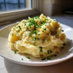 A generous serving of fluffy garlic mashed potatoes topped with freshly chopped green chives on a white plate.