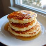 A tall stack of three fluffy old fashioned pancakes drizzled with maple syrup on a white plate.
