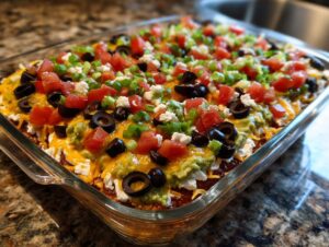 A fully assembled Seven layer dip topped with tomatoes, olives, cheese, and green onions in a clear glass baking dish.