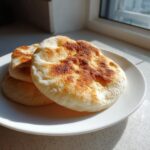 Three freshly cooked, slightly charred pita bread rounds stacked on a white plate near a sunlit window.