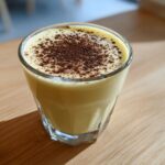 A close-up of a glass filled with creamy homemade eggnog, topped with a dusting of dark spice like nutmeg or cinnamon.