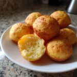 A pile of golden-brown Brazilian cheese bread puffs served on a white plate, one is broken open showing the airy interior.