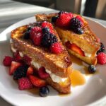 Two halves of golden stuffed french toast filled with cream and topped generously with fresh raspberries, blueberries, and blackberries.