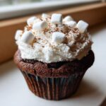 A close-up of a rich chocolate hot chocolate cupcake topped with whipped cream, mini marshmallows, and cocoa powder.