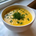 A close-up of a white bowl filled with creamy, yellow Italian penicillin soup, topped with fresh parsley.