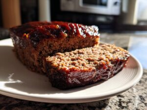 A thick slice cut from a glazed Keto meatloaf resting on a white plate in bright sunlight.