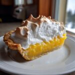 A tall slice of lemon meringue pie showing bright yellow filling and a thick, toasted meringue topping.