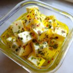 Close-up of cubed cheese soaking in bright yellow oil with herbs and red pepper flakes for a marinated cheese appetizer.