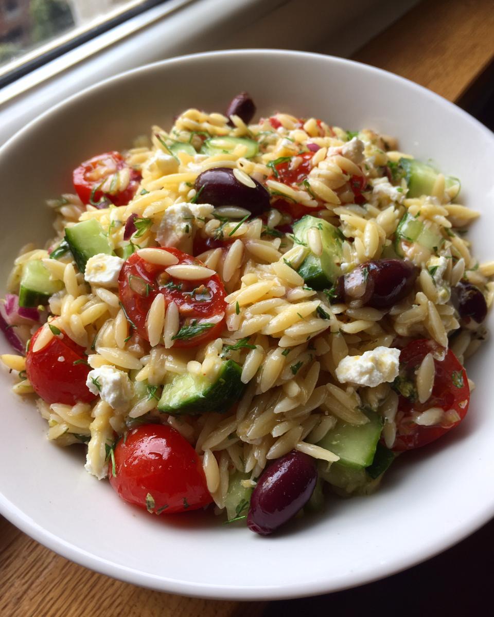 A bowl filled with vibrant mediterranean pasta salad featuring orzo, cherry tomatoes, cucumber, feta, and Kalamata olives.