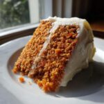A close-up, sunlit slice of moist carrot cake recipe with thick cream cheese frosting on a white plate.