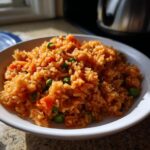 A close-up, appetizing photo of a white bowl filled with bright orange Spanish rice mixed with green peas and diced carrots.