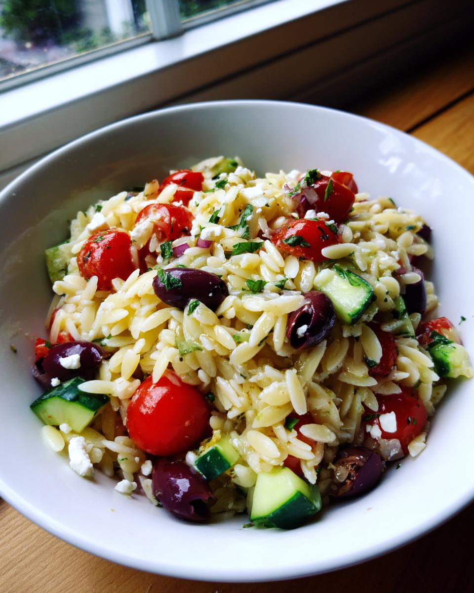 Close-up of a white bowl filled with mediterranean pasta salad featuring orzo, cherry tomatoes, cucumbers, olives, and feta.