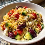 Close-up of a white bowl filled with mediterranean pasta salad made with orzo, tomatoes, cucumbers, olives, and feta.